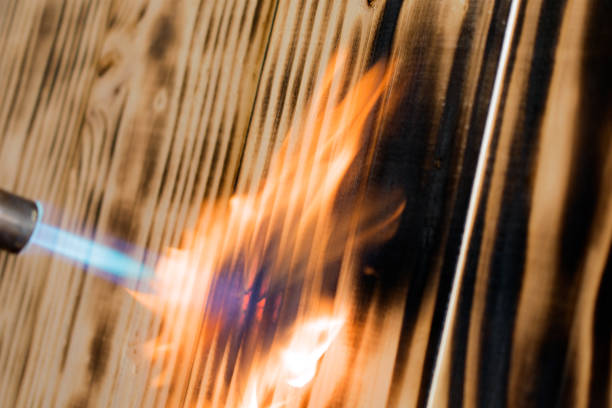 Ensayo de resistencia al fuego sobre tablero de madera con soplete mostrando combustión y carbonización superficial